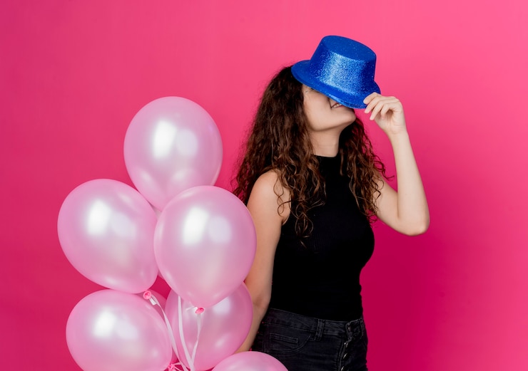 Foto gratuito giovane bella donna con capelli ricci in un cappello di festa che tiene il mazzo di mongolfiere in piedi sopra il muro rosa