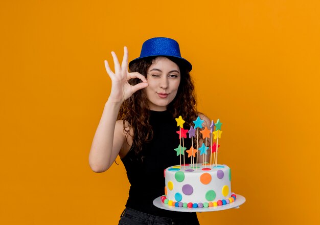 Giovane bella donna con capelli ricci in un cappello di festa che tiene la torta di compleanno che mostra segno giusto che sorride e che fa l'occhiolino in piedi sopra la parete arancione