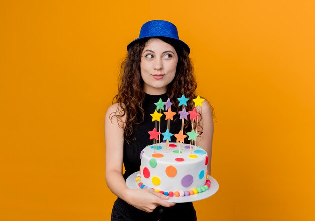 Giovane bella donna con capelli ricci in un cappello di festa che tiene la torta di compleanno che osserva da parte con il sorriso sul viso in piedi sopra la parete arancione