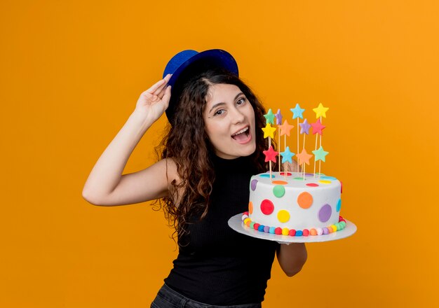 Giovane bella donna con capelli ricci in un cappello di festa che tiene la torta di compleanno felice e allegra in piedi sopra la parete arancione