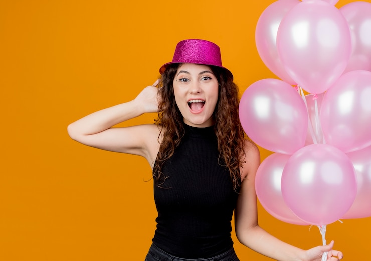 Foto gratuito giovane bella donna con capelli ricci in un cappello di festa che tiene mazzo di palloncini d'aria concetto di festa di compleanno felice ed eccitato in piedi sopra la parete arancione