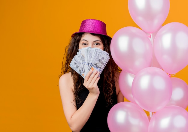 Giovane bella donna con capelli ricci in un cappello di festa che tiene un mazzo di mongolfiere che mostra il concetto di festa di compleanno felice ed eccitato in contanti in piedi sopra la parete arancione