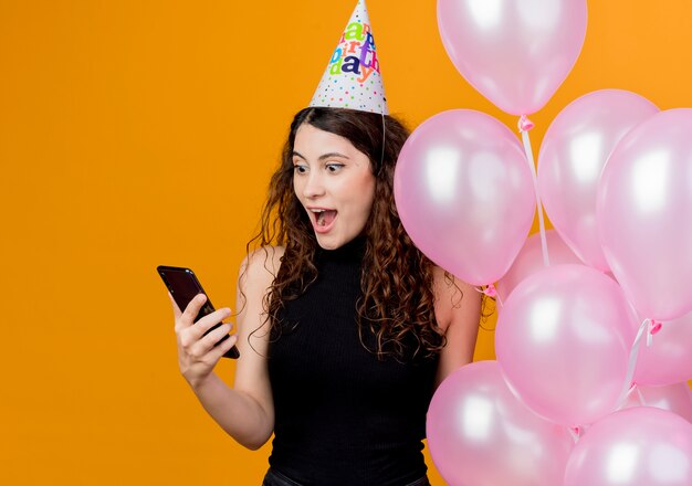 Giovane bella donna con capelli ricci in un cappello di vacanza che tiene gli aerostati di aria guardando lo schermo del suo concetto di festa di compleanno felice ed eccitato smartphone in piedi sopra la parete arancione