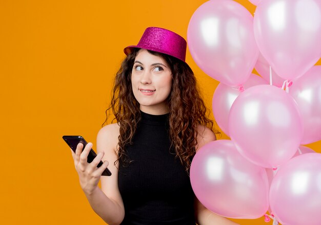 Giovane bella donna con capelli ricci in un cappello di vacanza che tiene il mazzo di aria palloncini e smartphone alzando lo sguardo pensando a mordere le labbra concetto di festa di compleanno in piedi sopra la parete arancione