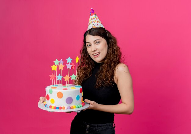 Giovane bella donna con capelli ricci in una protezione di festa che tiene il concetto di festa di compleanno felice e positivo della torta di compleanno che sta sopra la parete rosa