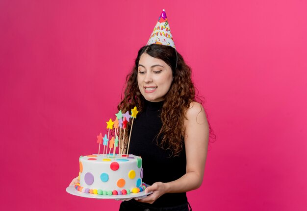 Giovane bella donna con capelli ricci in una protezione di festa che tiene il concetto di festa di compleanno felice e positivo della torta di compleanno che sta sopra la parete rosa