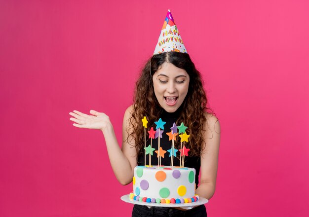 Giovane bella donna con capelli ricci in una protezione di festa che tiene il concetto di festa di compleanno felice ed emozionato della torta di compleanno che sta sopra la parete rosa