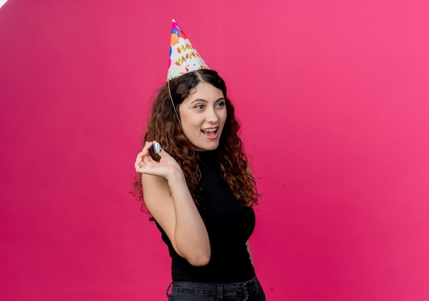 Giovane bella donna con capelli ricci in una protezione di festa che tiene il concetto felice e positivo della festa di compleanno del fischio che sta sopra la parete rosa
