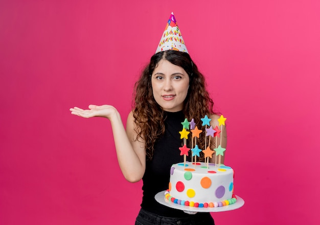 Giovane bella donna con capelli ricci in una protezione di festa che tiene la torta di compleanno che sembra confusa che scrolla le spalle concetto della festa di compleanno delle spalle che sta sopra la parete rosa