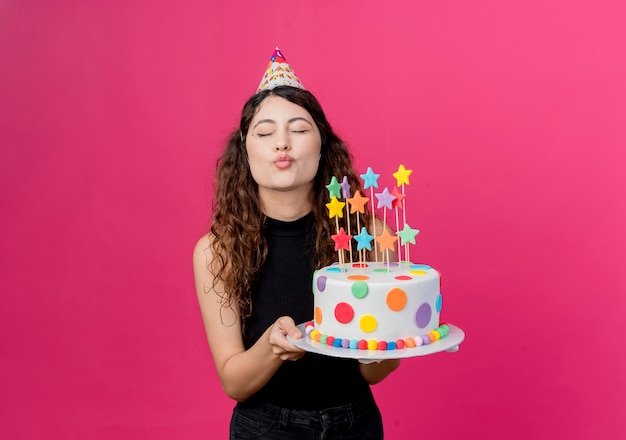 Giovane bella donna con capelli ricci in una protezione di festa che tiene la torta di compleanno che soffia un concetto di festa di compleanno di bacio che sta sopra la parete rosa