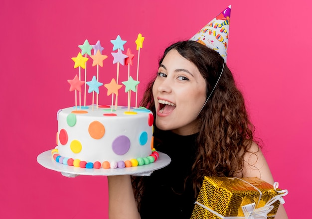 Giovane bella donna con capelli ricci in una protezione di festa che tiene la torta di compleanno e il concetto di festa di compleanno felice ed emozionato del contenitore di regalo sopra il rosa