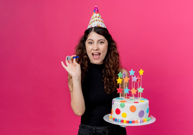 Giovane bella donna con capelli ricci in una protezione di festa che tiene la torta di compleanno felice ed emozionato che sorride allegramente concetto di festa di compleanno in piedi sopra il muro rosa