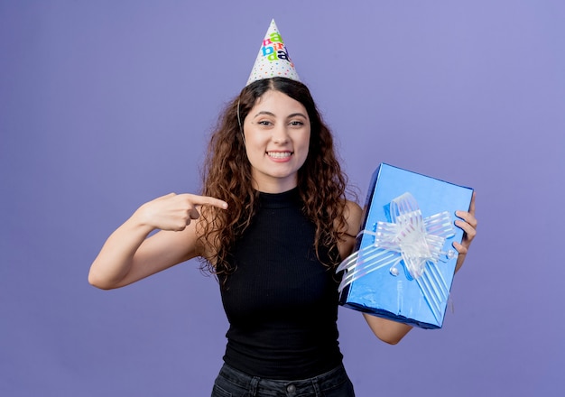 Giovane bella donna con capelli ricci in una protezione di vacanza che tiene il contenitore di regalo di compleanno che indica con il dito sorridendo allegramente concetto di festa di compleanno in piedi sopra la parete blu