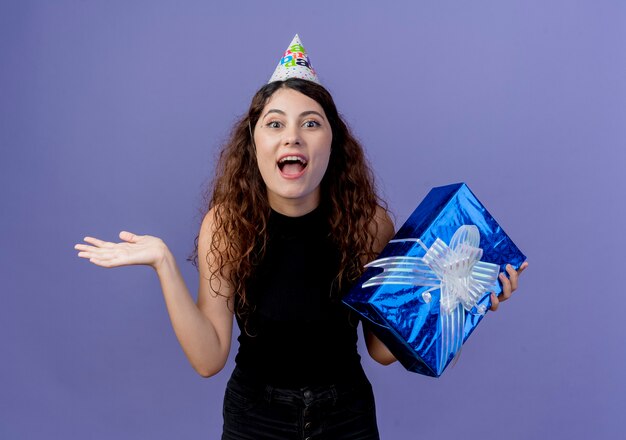 Giovane bella donna con capelli ricci in una protezione di vacanza che tiene il contenitore di regalo di compleanno che sembra confuso e sorpreso sorridente allegramente concetto di festa di compleanno in piedi sopra la parete blu