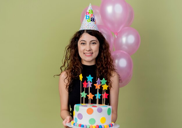 Giovane bella donna con capelli ricci in una protezione di vacanza che tiene la torta di compleanno che sorride allegramente felice e gioiosa in piedi sopra la parete chiara