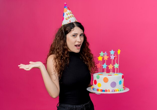 Giovane bella donna con capelli ricci in una protezione di vacanza che tiene la torta di compleanno confuso il concetto di festa di compleanno in piedi sopra la parete rosa