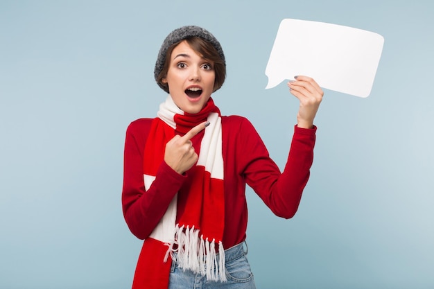 Giovane bella donna con i capelli corti scuri in maglione rosso, sciarpa e cappello lavorato a maglia guardando sorprendentemente nella fotocamera mentre tiene in mano la forma del messaggio di carta bianca su sfondo blu