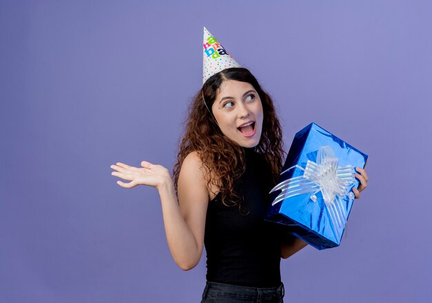 Giovane bella donna con i capelli ricci in un cappello di festa che tiene il contenitore di regalo di compleanno che sembra stupito e sorpreso sorridente allegramente concetto di festa di compleanno in piedi sopra la parete blu