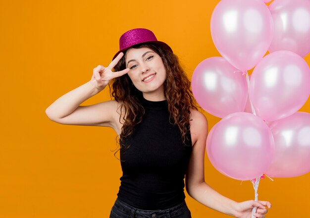 Giovane bella donna con i capelli ricci in un cappello di festa che tiene il mazzo di mongolfiere sorridente felice e positivo che mostra allegramente il concetto di festa di compleanno di v-segno in piedi sopra la parete arancione