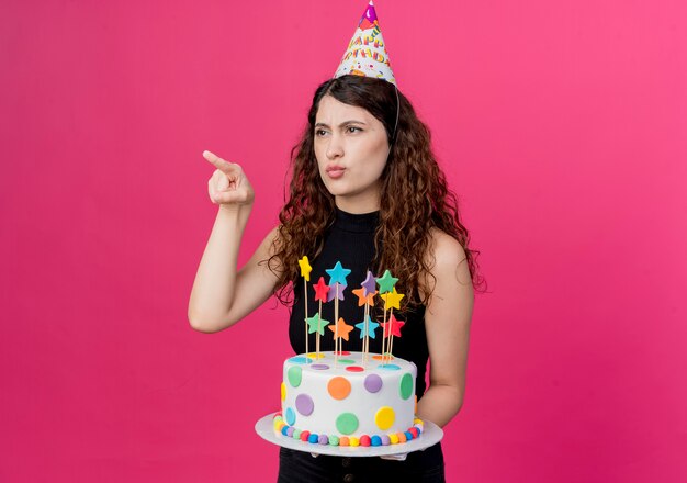 Giovane bella donna con i capelli ricci in un cappello di vacanza tenendo la torta di compleanno puntando il dito contro qualcosa che guarda il concetto di festa di compleanno scontento in piedi sopra il muro rosa