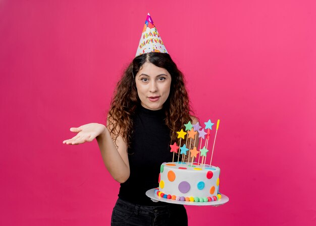 Giovane bella donna con i capelli ricci in un cappello di vacanza tenendo la torta di compleanno sorridente con il braccio fuori come chiedendo il concetto di festa di compleanno in piedi sopra la parete rosa