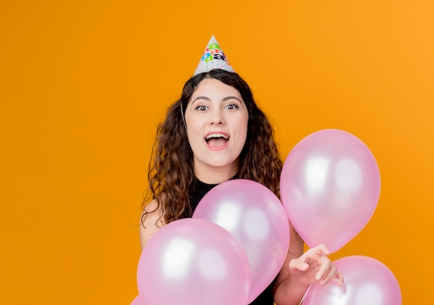 Giovane bella donna con i capelli ricci in una protezione di vacanza che tiene gli aerostati di aria happyand eccitato concetto di festa di compleanno in piedi sopra la parete arancione