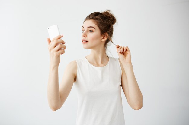 Giovane bella donna con il panino che esamina lo schermo del telefono che corregge capelli sopra fondo bianco.