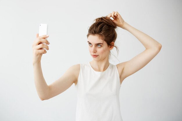 Giovane bella donna con il panino che esamina lo schermo del telefono che corregge capelli sopra fondo bianco.