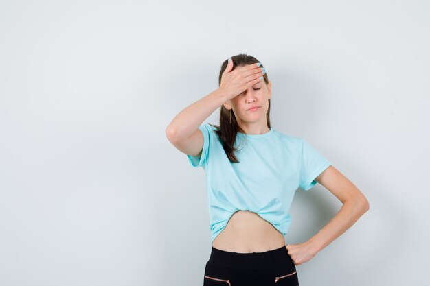 Giovane bella donna con le mani sulla fronte in t-shirt, pantaloni e guardando esausto, vista frontale.