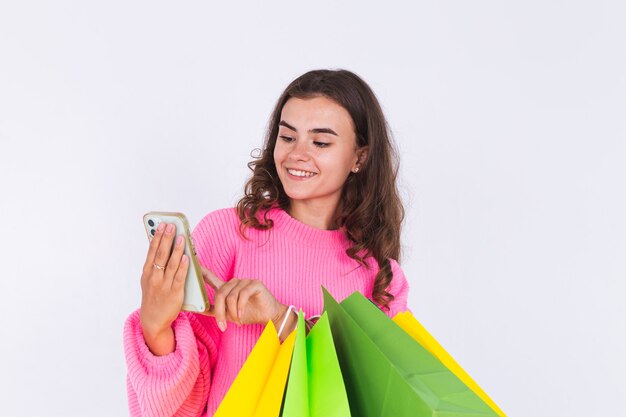 Giovane bella donna con lentiggini trucco leggero in maglione sul muro bianco con borse della spesa e telefono cellulare sorridente allegro positivo