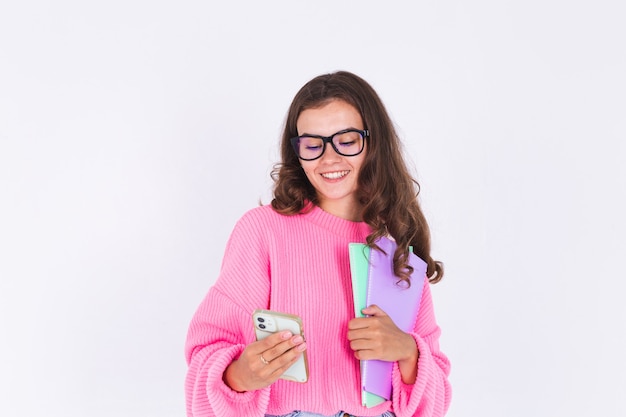 Giovane bella donna con lentiggini trucco leggero in maglione sul muro bianco studente con telefono cellulare sguardo premuroso allo schermo e sorriso