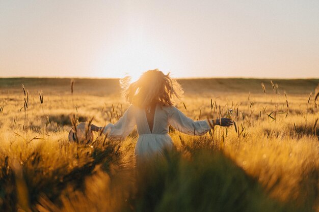 giovane bella donna con lunghi capelli biondi in un vestito bianco su un campo di grano al mattino presto all'alba. L'estate è tempo di sognatori, capelli svolazzanti, una donna che corre attraverso il campo sotto i raggi