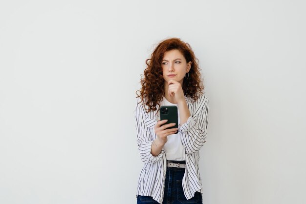 Giovane bella donna con lunghi capelli rossi naturali che pensa all'espressione facciale e posa che tiene il cellulare cellulare Attraente donna hipster premurosa con lo smartphone in mano