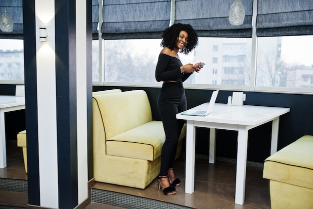 Giovane bella donna d'affari afroamericana alla moda con l'acconciatura afro in elegante supporto nero vicino al computer portatile e guardando sul telefono cellulare