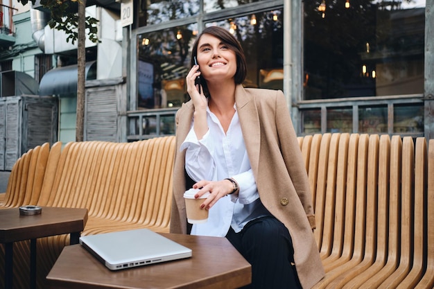 Giovane bella donna d'affari sorridente con il computer portatile che parla felicemente sul cellulare durante la pausa caffè nella caffetteria sulla strada