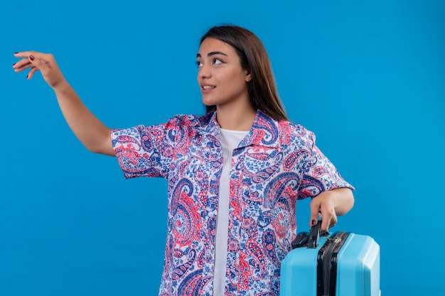 Giovane bella donna del viaggiatore che tiene la valigia blu che osserva da parte gesticolando con la mano che chiede di venire in piedi su sfondo blu