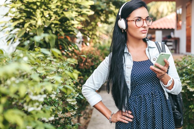 Giovane bella donna elegante che utilizza smartphone, cuffie, occhiali da vista, estate, vestito di denim vintage, sorridente, felice, positivo