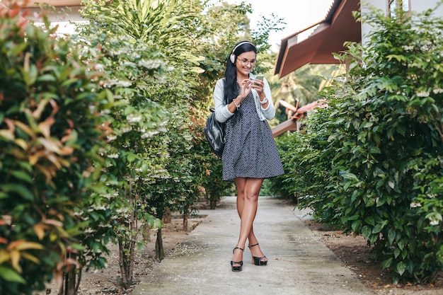 Giovane bella donna elegante che utilizza smartphone, cuffie, occhiali da vista, estate, vestito di denim vintage, sorridente, felice, positivo