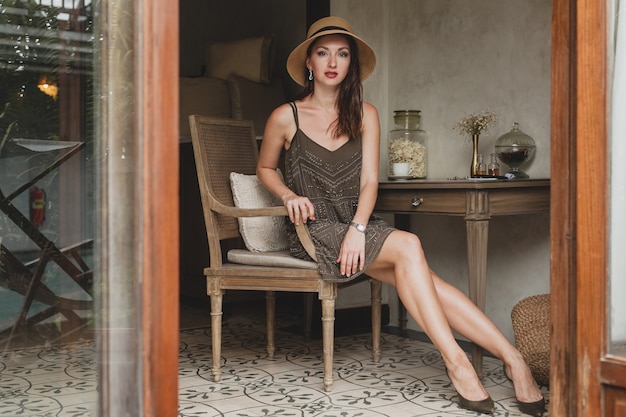 Giovane bella donna elegante nella camera d'albergo del resort, seduto al tavolo, indossando abiti alla moda, stile safari, cappello di paglia, sorridente, felice, vacanze estive, vestito bohémien