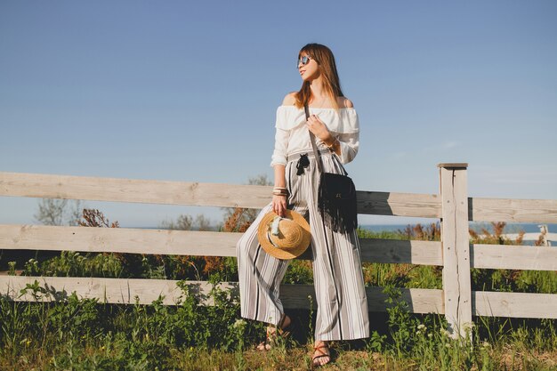 Giovane bella donna elegante, tendenza moda primavera estate, stile boho, cappello di paglia, weekend in campagna, sole, sorridente, divertimento, occhiali da sole, borsa nera, pantaloni a righe