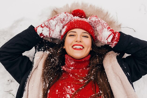 Giovane bella donna felice sorridente in guanti rossi e berretto lavorato a maglia che indossa cappotto invernale giacente nel parco nella neve, vestiti caldi, vista da sopra