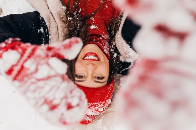 Giovane bella donna felice sorridente in guanti rossi e berretto lavorato a maglia che indossa cappotto invernale giacente nel parco nella neve, vestiti caldi, vista da sopra