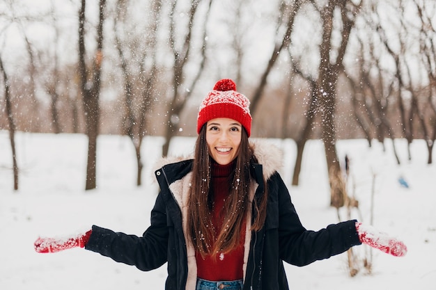 Giovane bella donna felice sorridente in guanti rossi e cappello lavorato a maglia che indossa cappotto invernale, passeggiate nel parco il giorno di nevicata, vestiti caldi