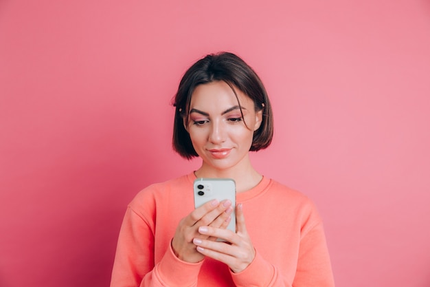 Giovane bella donna felice sveglia che posa isolata sopra il fondo rosa della parete facendo uso del telefono cellulare