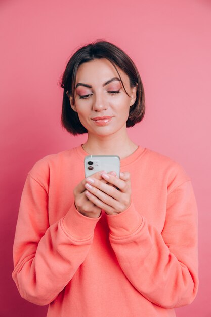 Giovane bella donna felice sveglia che posa isolata sopra il fondo rosa della parete facendo uso del telefono cellulare