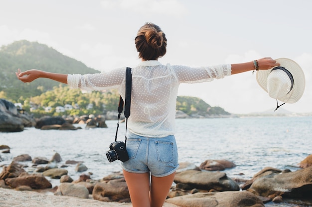 Giovane bella donna hipster in vacanza estiva in asia, rilassante sulla spiaggia tropicale, fotocamera digitale, stile boho casual, paesaggio marino, corpo abbronzato sottile, viaggio da solo, libertà