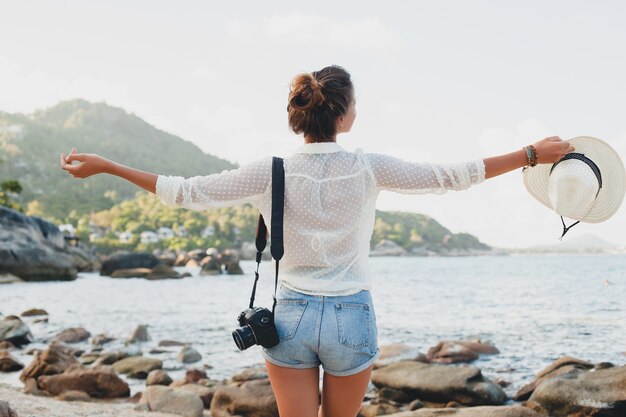 Giovane bella donna hipster in vacanza estiva in asia, rilassante sulla spiaggia tropicale, fotocamera digitale, stile boho casual, paesaggio marino, corpo abbronzato sottile, viaggio da solo, libertà