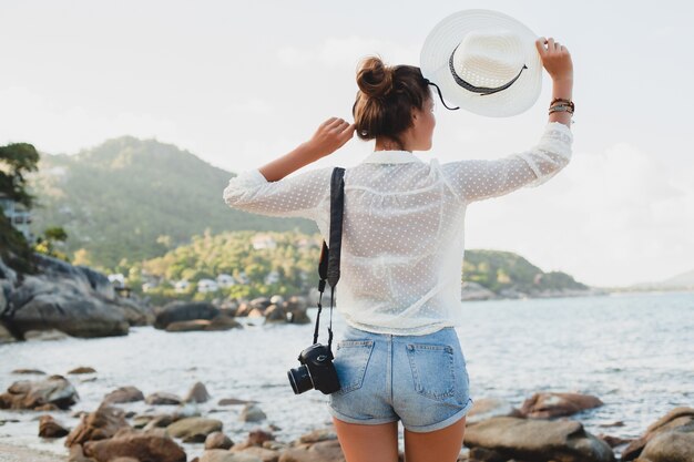 Giovane bella donna hipster in vacanza estiva in asia, rilassante sulla spiaggia tropicale, fotocamera digitale, stile boho casual, paesaggio marino, corpo abbronzato sottile, viaggio da solo, libertà