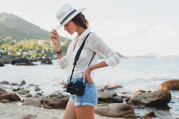 Giovane bella donna hipster in vacanza estiva in asia, rilassante sulla spiaggia tropicale, fotocamera digitale, stile boho casual, paesaggio marino, corpo abbronzato sottile, viaggio da solo