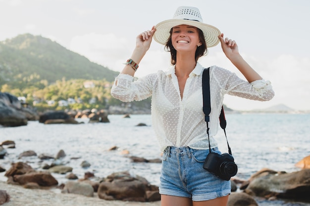 Giovane bella donna hipster in vacanza estiva in asia, rilassante sulla spiaggia tropicale, fotocamera digitale, stile boho casual, paesaggio marino, corpo abbronzato sottile, viaggio da solo